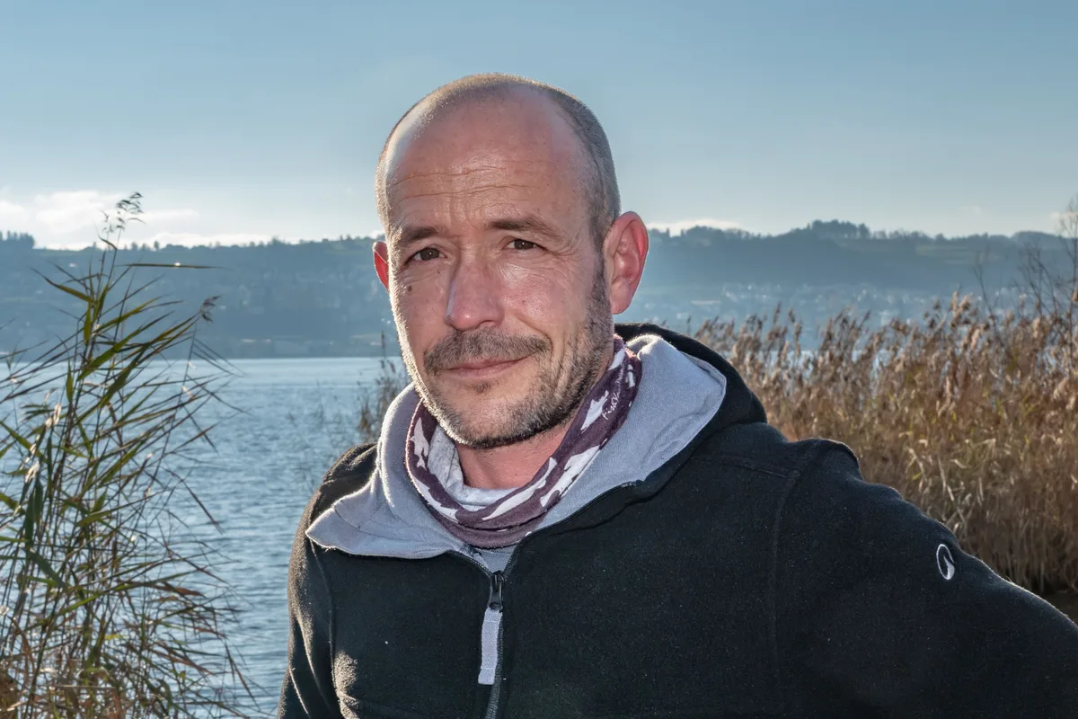 «Wir alle haben Angst»: Die Belastung der Gewässer mit PFAS und anderen Chemikalien beunruhigt die Schweizer Berufsfischer. Porträt von Berufsfischer Samuel Weidmann. Ein Mann mit sehr kurzem Haar und dunklem Hoodie steht am Zürichsee. Im Hintergrund ist Schilf zu sehen.